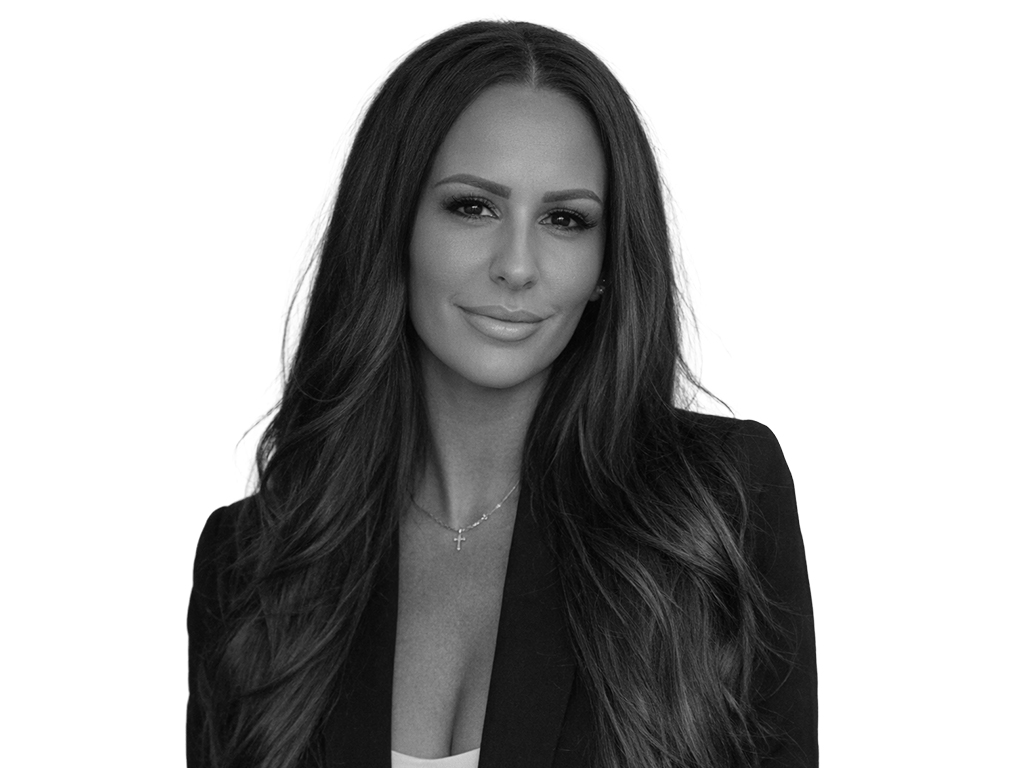 Black-and-white headshot of a woman with long wavy hair, wearing a blazer and a subtle necklace, looking at the camera with a soft smile.”] ,