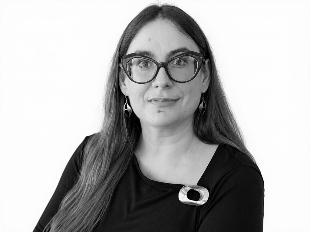 Portrait of a woman with long hair, large glasses, and drop earrings, facing the camera in a black top with a metallic pin on the shoulder.