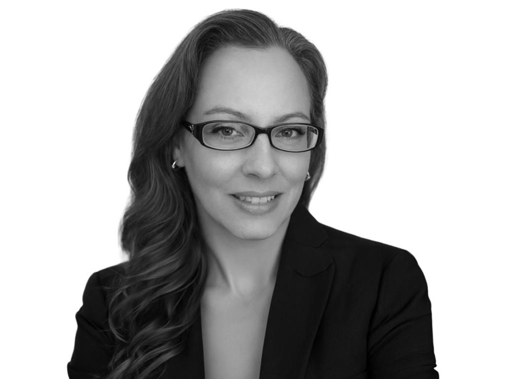 Professional headshot of a woman with dark hair, glasses, and a black blazer, smiling softly against a white background.