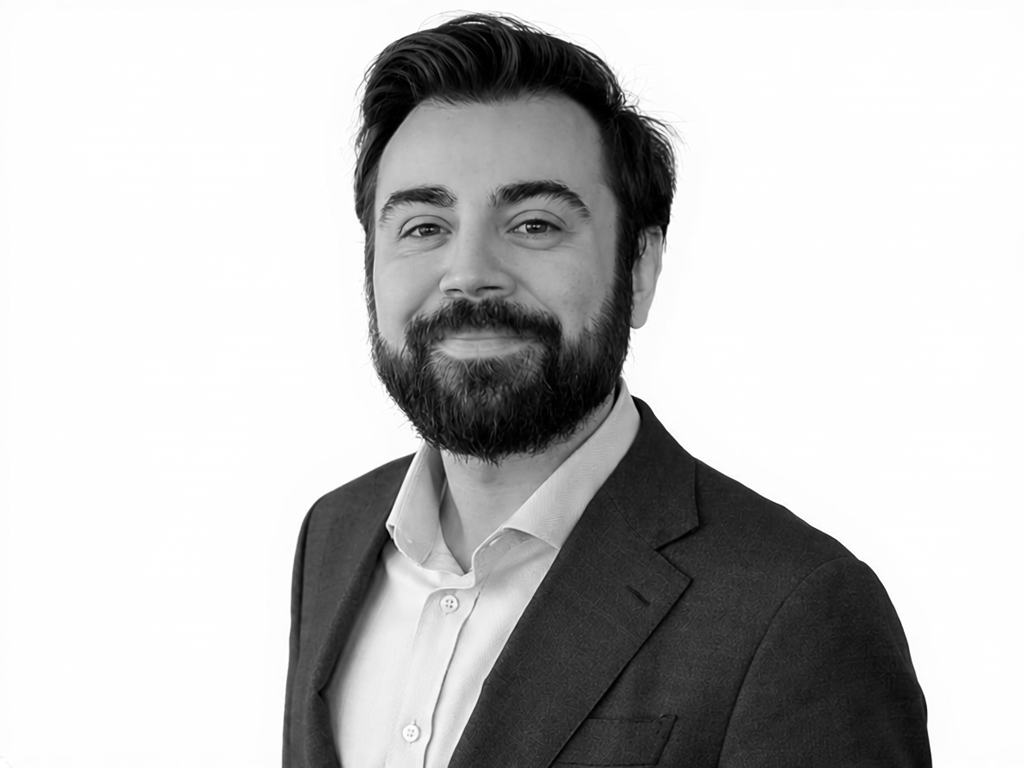 Professional headshot: man with dark hair and beard in a blazer and white shirt, smiling slightly against a light background